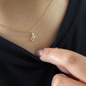 Op de afbeelding: Een gouden ketting met een hanger in de vorm van een cursieve letter "A". De ketting wordt gedragen op een zwarte top. De hanger hangt aan een fijne ketting en presenteert een eenvoudig en elegant ontwerp.