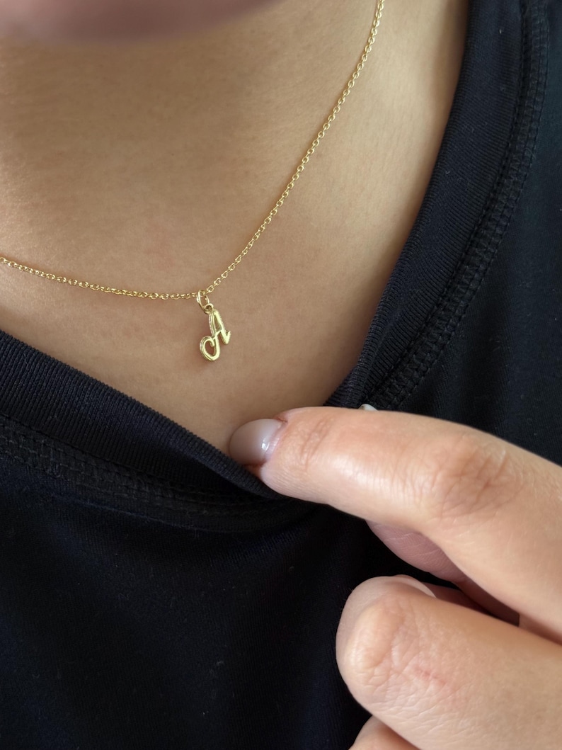 Op de afbeelding: Een gouden ketting met een hanger in de vorm van een cursieve letter "A". De ketting wordt gedragen op een zwarte top. De hanger hangt aan een fijne ketting en presenteert een eenvoudig en elegant ontwerp.