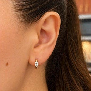Puede incluir: Primer plano de un pendiente de aro dorado con un solo diamante de forma ovalada. El pendiente es pequeño y delicado, con un diseño sencillo. El pendiente se lleva en la oreja de una persona.