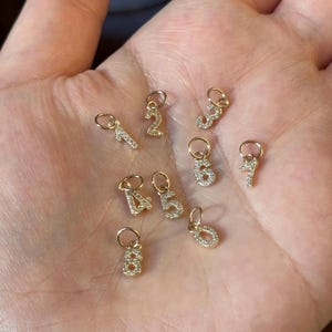 May include: A selection of small, gold-coloured number charms, each embellished with sparkling clear crystals. The numbers 1 to 8 are visible, each with a small loop for attachment. The charms are displayed against a neutral background.