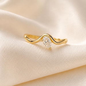 May include: A gold ring featuring a single pear-shaped clear gemstone set into a smooth, wavy band. The metal has a polished finish, and the stone is positioned at the peak of the curved design, creating a continuous, flowing silhouette.