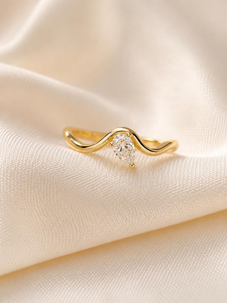May include: A gold ring featuring a single pear-shaped clear gemstone set into a smooth, wavy band. The metal has a polished finish, and the stone is positioned at the peak of the curved design, creating a continuous, flowing silhouette.