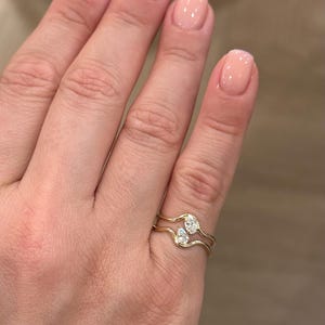 May include: A gold ring featuring two wavy bands that meet in the centre, each set with a small pear-shaped clear gemstone. The design creates a bypass style where the two stones are positioned vertically, one above the other, on the finger.