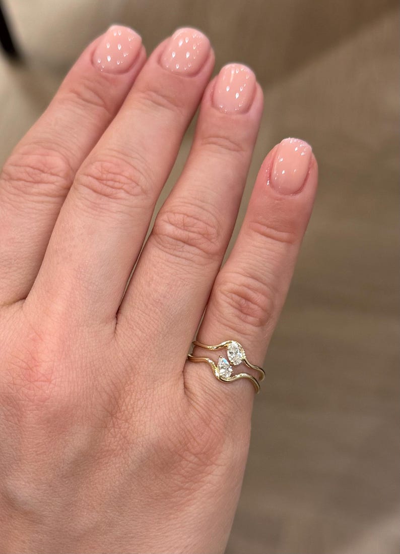 May include: A gold ring featuring two wavy bands that meet in the centre, each set with a small pear-shaped clear gemstone. The design creates a bypass style where the two stones are positioned vertically, one above the other, on the finger.