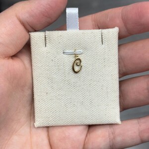 Op de afbeelding: Een goudkleurige letter "C" bedel, opgehangen aan een klein wit lint. De bedel wordt getoond op een beige stoffen kaart, gepresenteerd in een hand. De bedel is waarschijnlijk voor een ketting of armband.