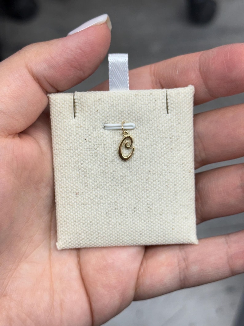 Op de afbeelding: Een goudkleurige letter "C" bedel, opgehangen aan een klein wit lint. De bedel wordt getoond op een beige stoffen kaart, gepresenteerd in een hand. De bedel is waarschijnlijk voor een ketting of armband.