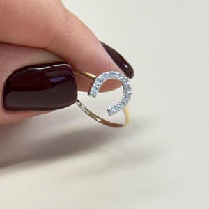 Op de afbeelding: Een gouden ring met een hoefijzerontwerp, bezet met kleine, heldere edelstenen. De ring wordt vastgehouden door een hand met donkerrode nagellak, die de delicate details en de glans van de ring benadrukt. Het hoefijzer is een symbool van geluk.