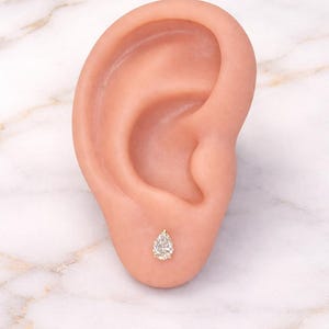 Può includere: Un orecchino a lobo a forma di goccia di diamante incastonato in oro. L'orecchino è mostrato su un orecchio umano, evidenziando le sue dimensioni e il suo design. Lo sfondo è una superficie marmorea.