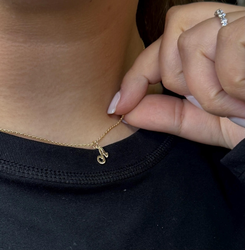 Op de afbeelding: Een delicate gouden ketting met een hanger in de vorm van een cursieve letter "A". De hanger hangt aan een fijne gouden ketting. De ketting wordt getoond tegen een zwarte achtergrond.
