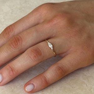 Può includere: Un sottile anello in oro con un diamante centrale taglio marquise affiancato da due diamanti più piccoli taglio rotondo. La fascia è sottile e lucida, caratterizzata da un design delicato che si posiziona orizzontalmente sul dito con tre pietre chiare.