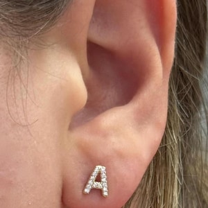 Könnte beinhalten: Nahaufnahme eines Ohrs mit einem kleinen, goldfarbenen Ohrstecker in Form des Buchstabens "A". Der Buchstabe ist mit kleinen, klaren Edelsteinen besetzt.