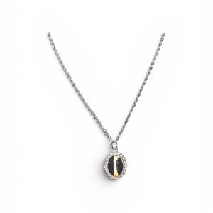 May include: Silver-toned necklace with an oval pendant. The pendant features a black background with a vertical yellow design and is framed by a ring of small, clear stones. The chain is delicate and has a textured appearance.