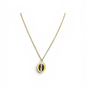 May include: A gold-colored necklace with a delicate chain and a pendant. The pendant is an oval shape with a black background and a white letter "I" in the center. The pendant is framed with a textured gold border.
