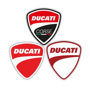 Puede incluir: Tres pegatinas con el logotipo de Ducati. La pegatina superior tiene forma de escudo con la palabra "DUCATI" en blanco sobre fondo rojo y "CORSE" sobre fondo negro. Las otras dos pegatinas son escudos rojos y blancos con la palabra "DUCATI".