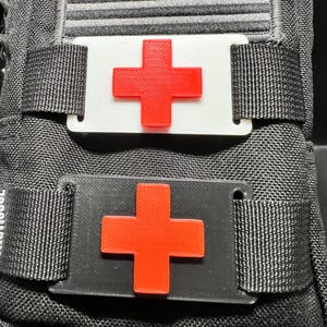 May include: Close-up of two medical patches with a red cross design. One patch is white, the other is black. Both patches are attached to a black fabric with straps.