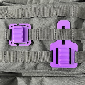 May include: Two purple MOLLE clips are shown, one open and one closed, against a backdrop of black webbing. The clips have a rectangular design with a series of horizontal ridges. The open clip has a C-shaped top.