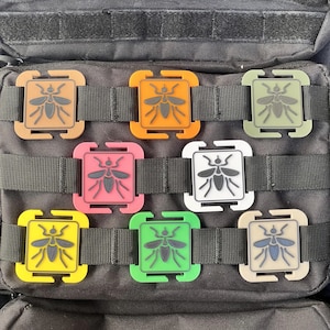 以下が含まれることがあります： スタイリッシュな昆虫のデザインが施された、7つのカラフルな正方形のパッチのコレクション。オレンジ、ピンク、緑、黄色など、さまざまな色があり、グリッドパターンを持つ黒色のテクスチャード加工された表面に取り付けられています。