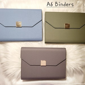 May include: Three A6 binders in different colors: light blue, dark green, and gray. Each binder has a gold metal closure.