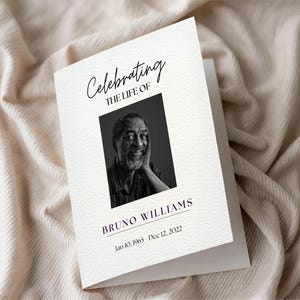 Op de afbeelding: Een gevouwen herdenkingskaart met de tekst "Celebrating the Life of" boven een zwart-witfoto van een man. De naam "Bruno Williams" en de data "Jan 10, 1965 - Dec 12, 2022" staan eronder gedrukt. De kaart ligt op een crèmekleurige stof.