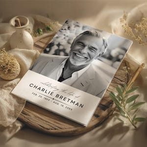 May include: A memorial card with a black and white photo of a smiling man. The card reads "celebrating the life of CHARLIE BRETMAN" with dates. The card is on a wooden tray with decorative elements.