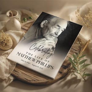 Peut inclure: Une carte commémorative avec un portrait en noir et blanc d'un homme. La carte porte l'inscription "Célébrer la vie de Matthew Phillips" avec ses dates de naissance et de décès. Elle est posée sur un plateau en bois.