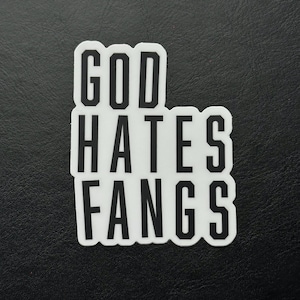 May include: A white sticker with the text "GOD HATES FANGS" in a bold, black font. The sticker has a rounded rectangular shape and is set against a dark background.