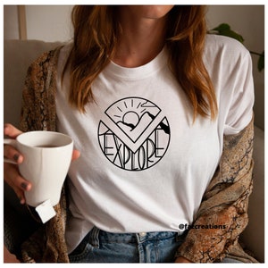 May include: White t-shirt with a black graphic design of a sun, mountains, and the word "explore" in a circle.