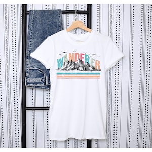 May include: White t-shirt with a graphic of mountains and the word "WANDERER" in colorful letters. The graphic has a vintage look with a blue, green, and orange color scheme.