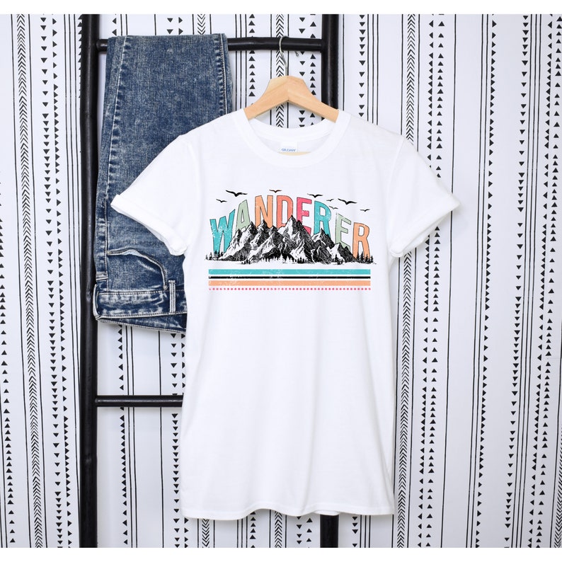 May include: White t-shirt with a graphic of mountains and the word "WANDERER" in colorful letters. The graphic has a vintage look with a blue, green, and orange color scheme.