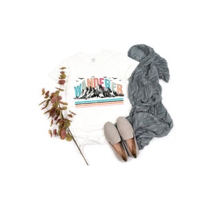 May include: White t-shirt with a graphic of mountains and the word "WANDERER" in colorful letters. The shirt is laid flat on a white background with a gray scarf and brown shoes.