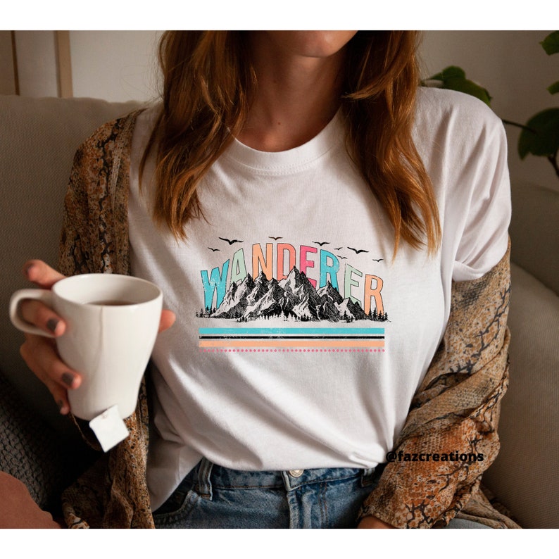 May include: White t-shirt with a graphic design of mountains and the word "WANDERER" in rainbow colors. The graphic has a vintage look with a faded effect.