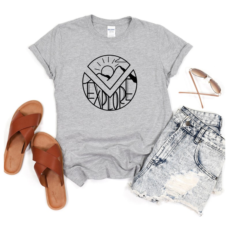 May include: A gray t-shirt with a black graphic of a sun, mountains, and the word "explore" in a circle. The shirt is laid flat on a white background with a pair of brown sandals and denim shorts.