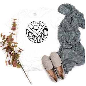 May include: White t-shirt with a black graphic design of a sun, mountains, and the word "explore" in a circle.