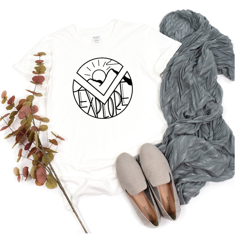May include: White t-shirt with a black graphic design of a sun, mountains, and the word "explore" in a circle.