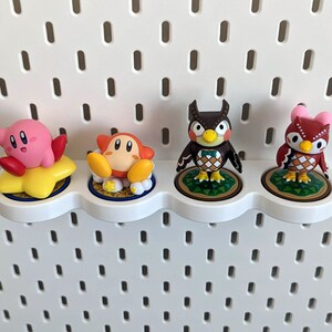 May include: Four character figurines on a white pegboard shelf. The figurines are of a pink and red Kirby, a yellow and orange character with a star, a brown owl with a green bow tie, and a red owl with pink ears.