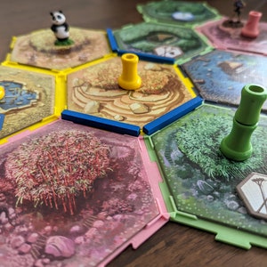 May include: A close-up of a hexagonal game board with colorful tiles and wooden tokens. The board features a panda figurine, bamboo, and a variety of other nature-themed illustrations. The tokens are in different colors, including yellow, green, and pink.
