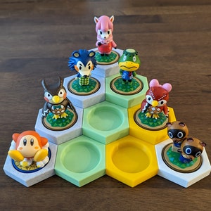 May include: A hexagonal display stand with six compartments, each featuring a different Animal Crossing character figurine. The characters include a pink sheep, a blue wolf, a green frog, a brown owl, a red bird, and two brown dogs. The stand is made of white, green, and yellow plastic.