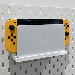 Könnte beinhalten: Ein weißes Wandregal mit einem gelb-schwarzen Nintendo Switch-Controller. Das Regal trägt die Aufschrift "Nintendo Switch".