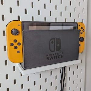 Puede incluir: Una consola Nintendo Switch negra con mandos Joy-Con amarillos montados en una pared de tablero de clavijas blanco. La consola está enchufada y tiene un estante blanco debajo.