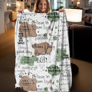 Könnte beinhalten: Weiße Kuscheldecke mit grünem und braunem Text und Illustrationen. Der Text enthält die Sätze "Olivia's Daily Reminders", "Focus!" und "I believe in me, I am loved".