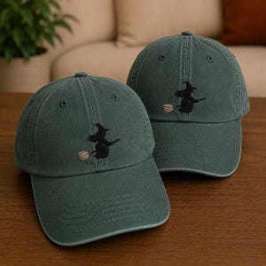 May include: Two dark green baseball caps featuring an embroidered black silhouette of a witch flying on a broomstick. The caps have a structured crown, a curved brim, and matching green eyelets, showcasing a simple and clean design aesthetic.