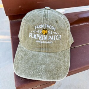 Puede incluir: Una gorra de b&eacute;isbol verde con el texto "Farm Fresh Pumpkin Patch pick your own" bordado en blanco. Una peque&ntilde;a calabaza naranja est&aacute; bordada en el centro del texto.