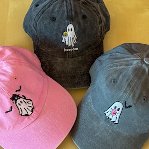 以下が含まれることがあります： ピンク、グレー、濃いグレーの野球帽3つ。各キャップには、刺繍された幽霊のデザインが施されています。濃いグレーのキャップには、飲み物を持っている幽霊と下に「boooze」という言葉があります。ピンクのキャップにはコウモリの幽霊があり、グレーのキャップにはピンクの舌の幽霊があります。