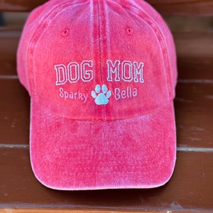 May include: Red baseball cap with the text "DOG MOM" embroidered in white. The names "Sparky" and "Bella" are embroidered below the text with a paw print in between.