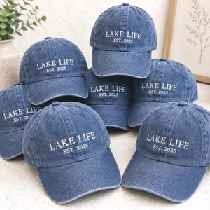 Könnte beinhalten: Eine Sammlung von Baseballkappen aus blauem Denim mit weißem Sticktext, der LAKE LIFE EST. 2025 lautet. Jede Kappe hat einen gebogenen Schirm, eine strukturierte Krone und ein klassisches Sechs-Panel-Design mit einem Knopf an der Oberseite.