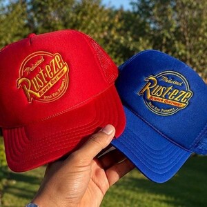May include: A red trucker hat and a blue trucker hat, both featuring an embroidered circular logo that reads Medicated Rust-eze Bumper Ointment. Each hat has a solid front panel, a mesh back, and a curved brim, showcasing a classic design.