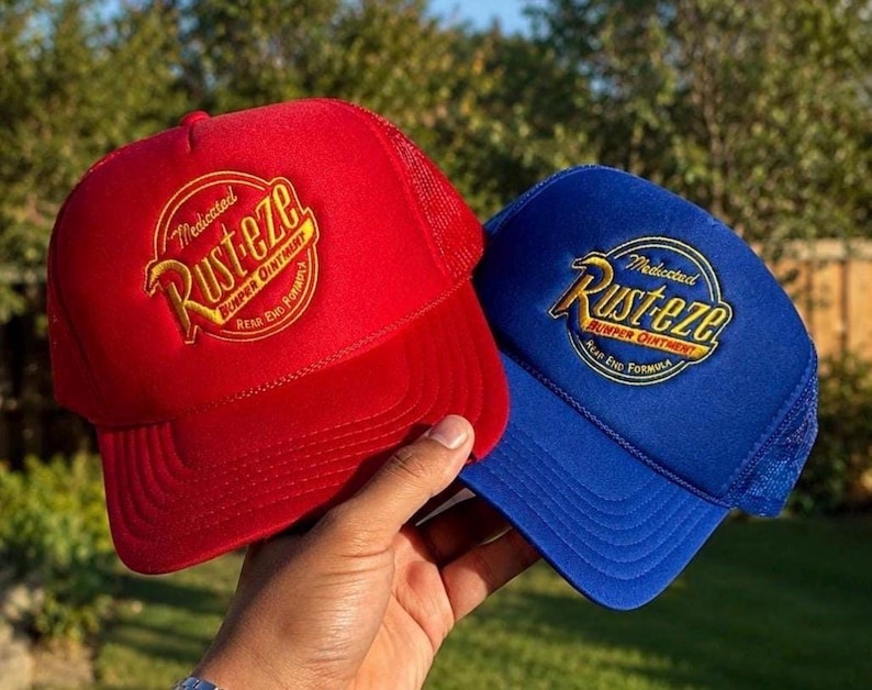 May include: A red trucker hat and a blue trucker hat, both featuring an embroidered circular logo that reads Medicated Rust-eze Bumper Ointment. Each hat has a solid front panel, a mesh back, and a curved brim, showcasing a classic design.