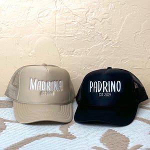 May include: Two trucker hats, one beige and one black, with white embroidered text. The beige hat says "Madrina Est. 2024" and the black hat says "Padrino Est. 2024".