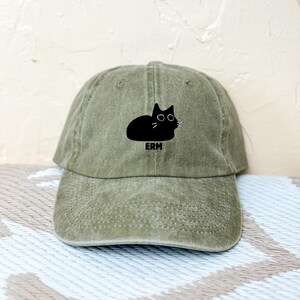 Puede incluir: Una gorra de béisbol verde con una ilustración de un gato negro y la palabra "ERM" impresa en la parte delantera.