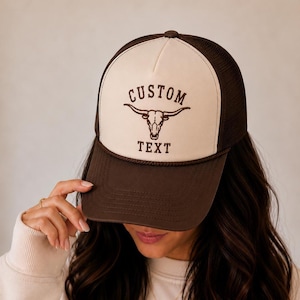 May include: A brown and cream trucker hat featuring a dark brown mesh back and a solid cream front panel. The front displays a dark brown graphic of a longhorn skull with the word CUSTOM arched above and the word TEXT printed below the skull.